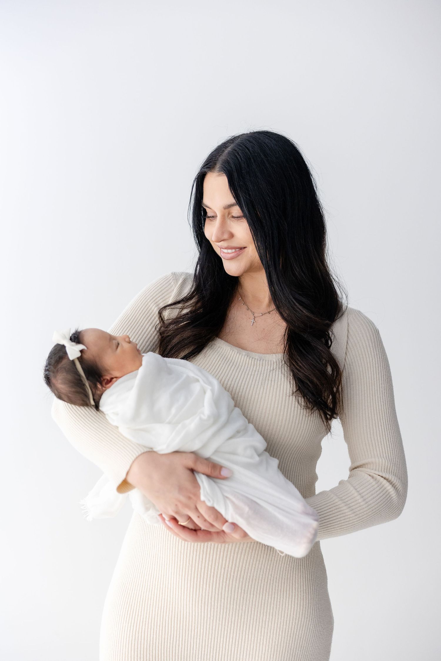 Woman in beige ribbed dress holds newborn baby wrapped in white blanket against white background.