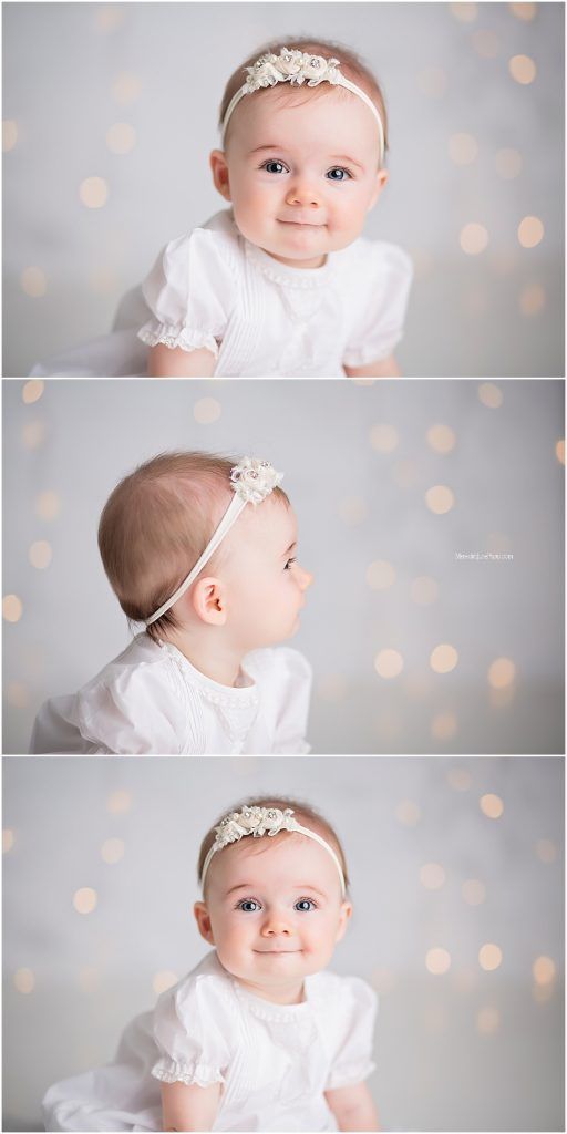 Baby in white dress with delicate headband poses against soft bokeh background for professional portrait session.
