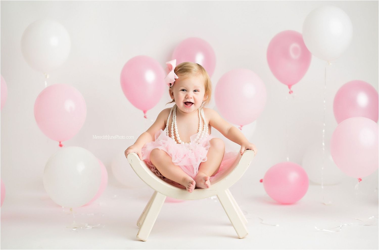 Baby in pink dress poses with pink balloons during first birthday photo shoot.