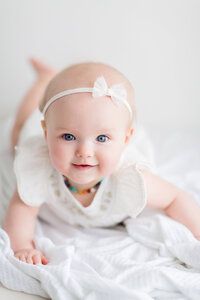 A baby wearing white clothing lies on white bedding in a bright, airy setting.