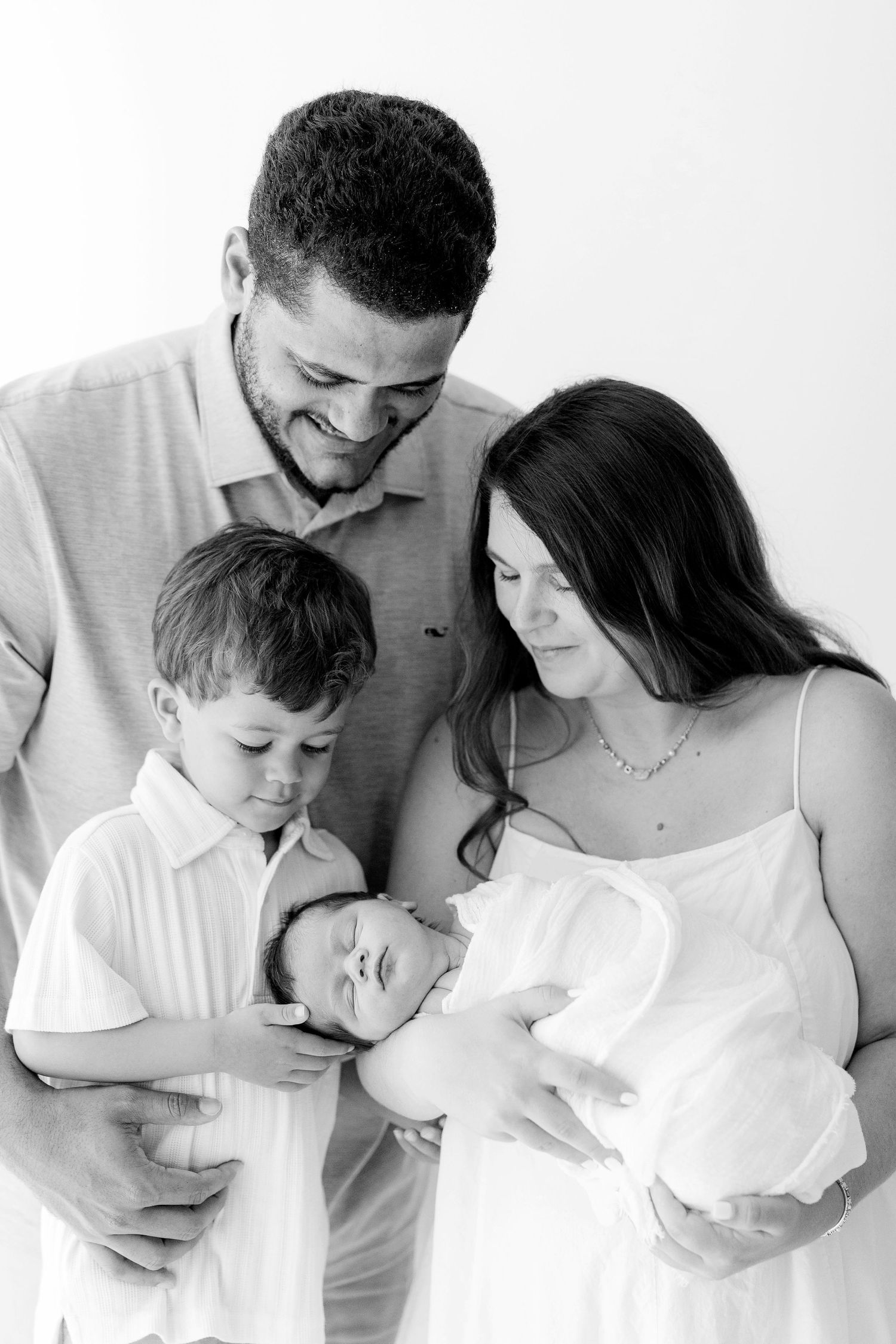 Black and white family portrait of parents and young son lovingly gazing at newborn baby in mothers arms.