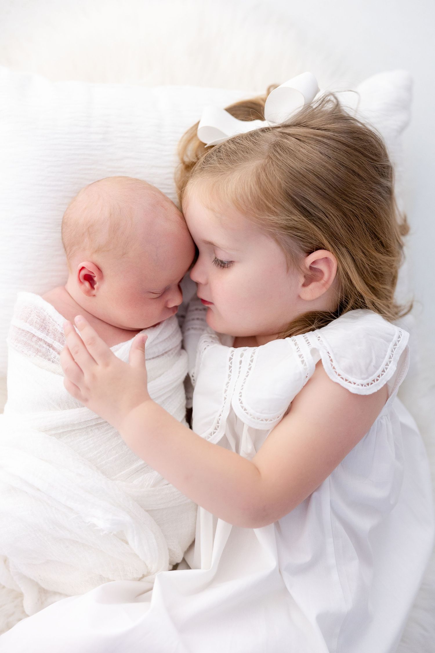 Young girl in white dress tenderly cuddles newborn baby sibling on white bedding.