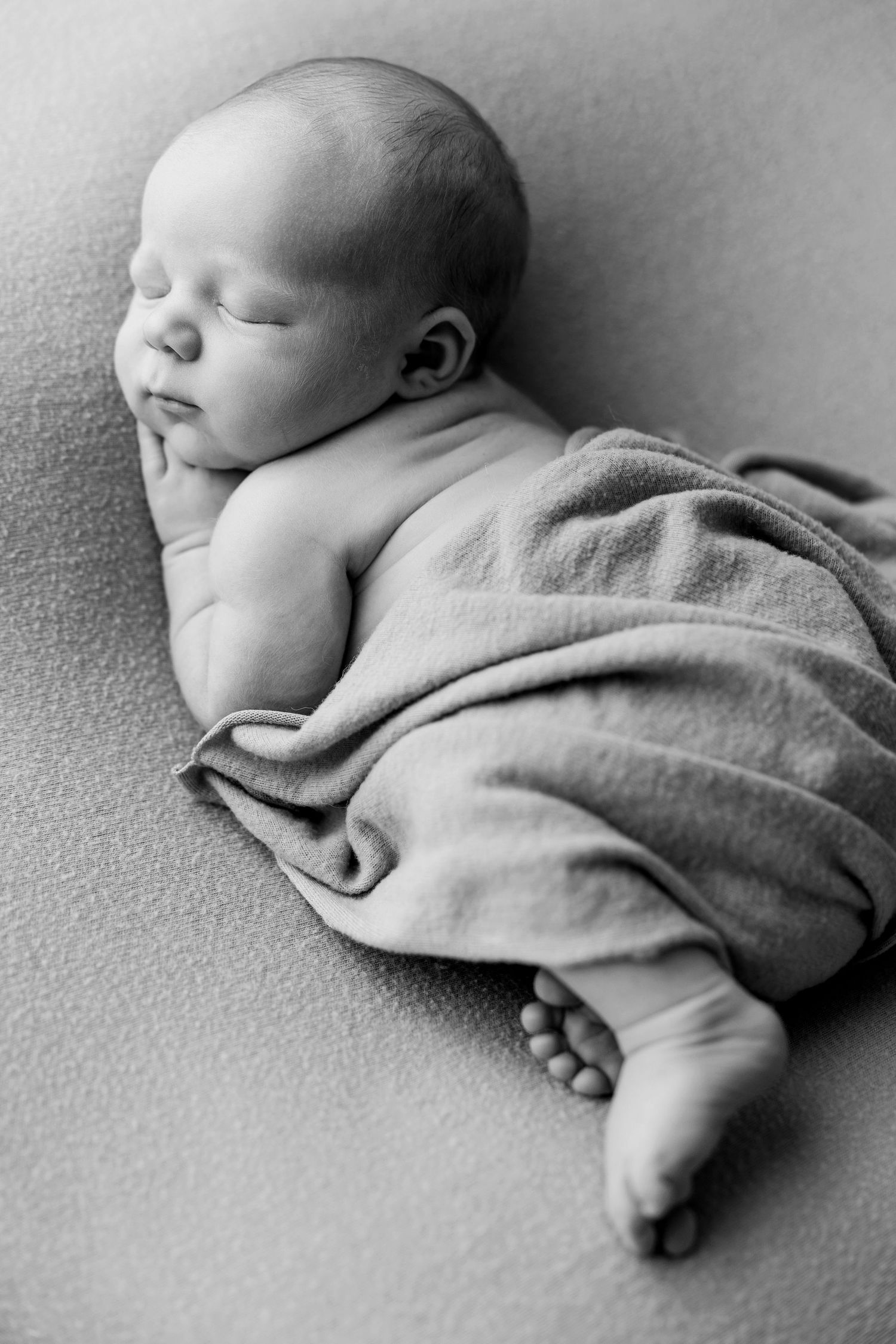 Black and white photo of a peaceful sleeping newborn baby wrapped in a soft blanket with tiny toes visible.
