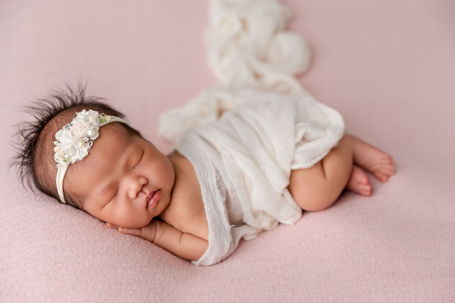 Peaceful newborn baby girl sleeping wrapped in white lace on a pink blanket with a floral headband.