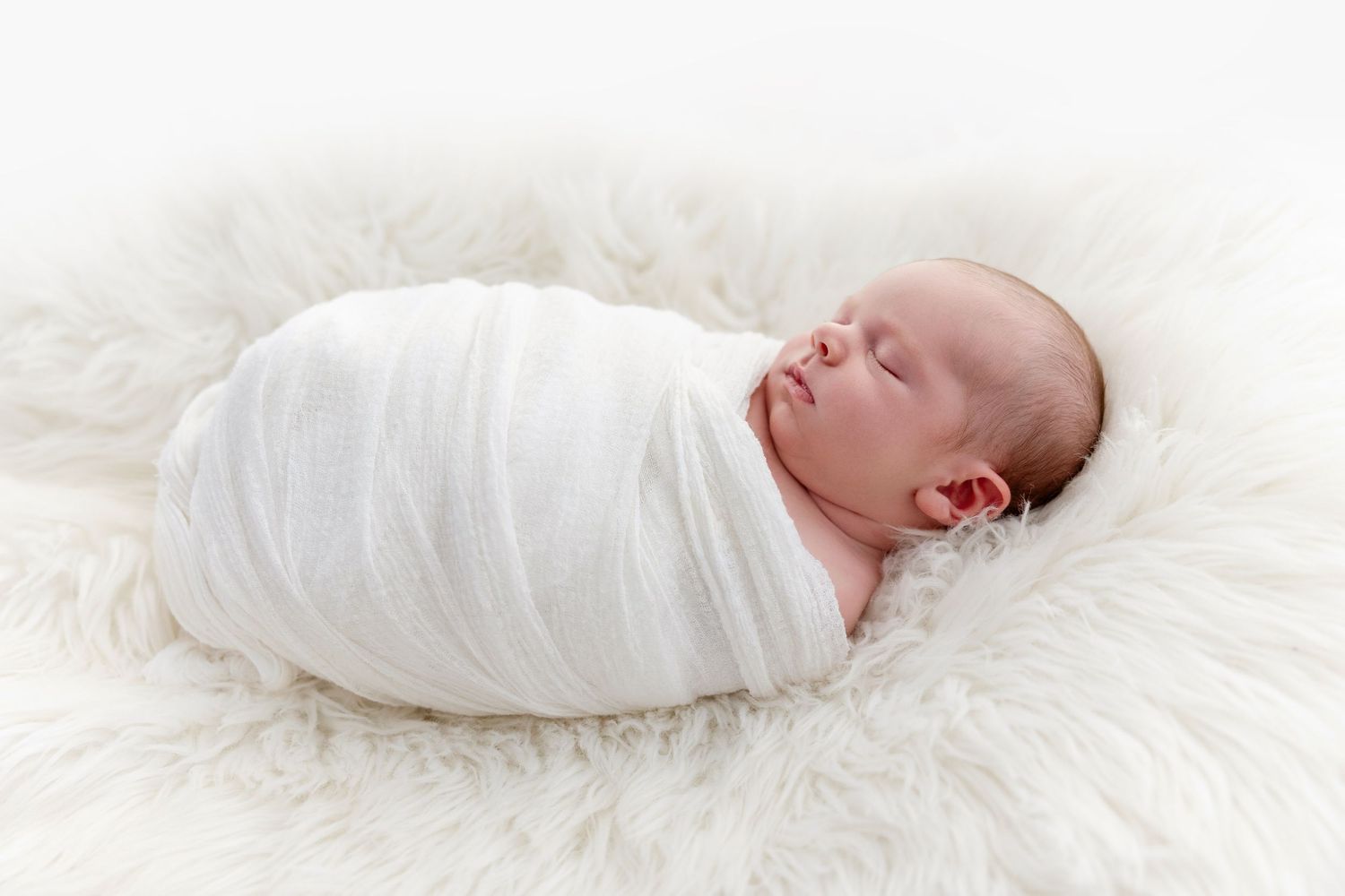Newborn baby swaddled in white wrap sleeping peacefully on a fluffy white fur rug.