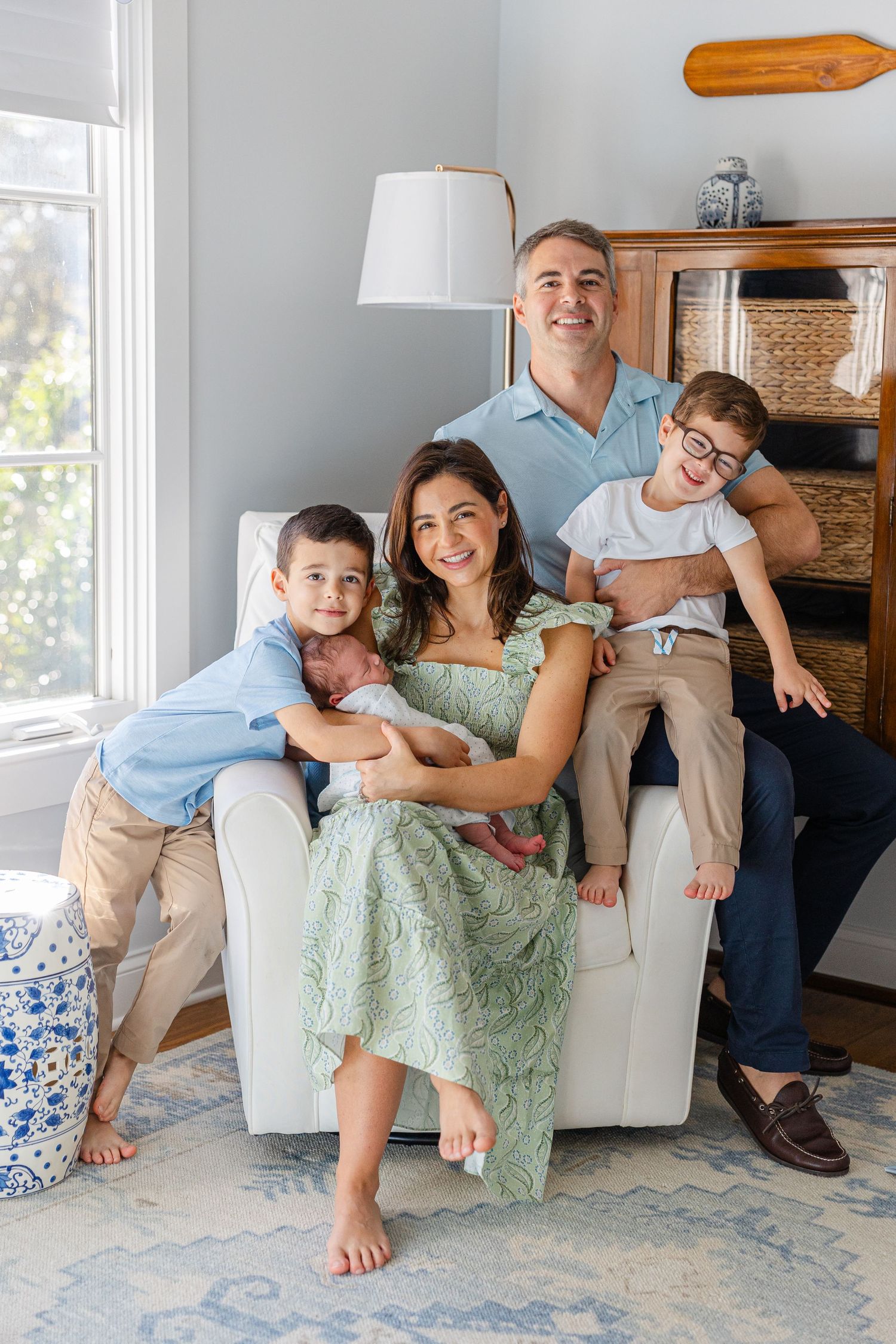 Family relaxes together on white couch in bright living room with blue rug and natural lighting.