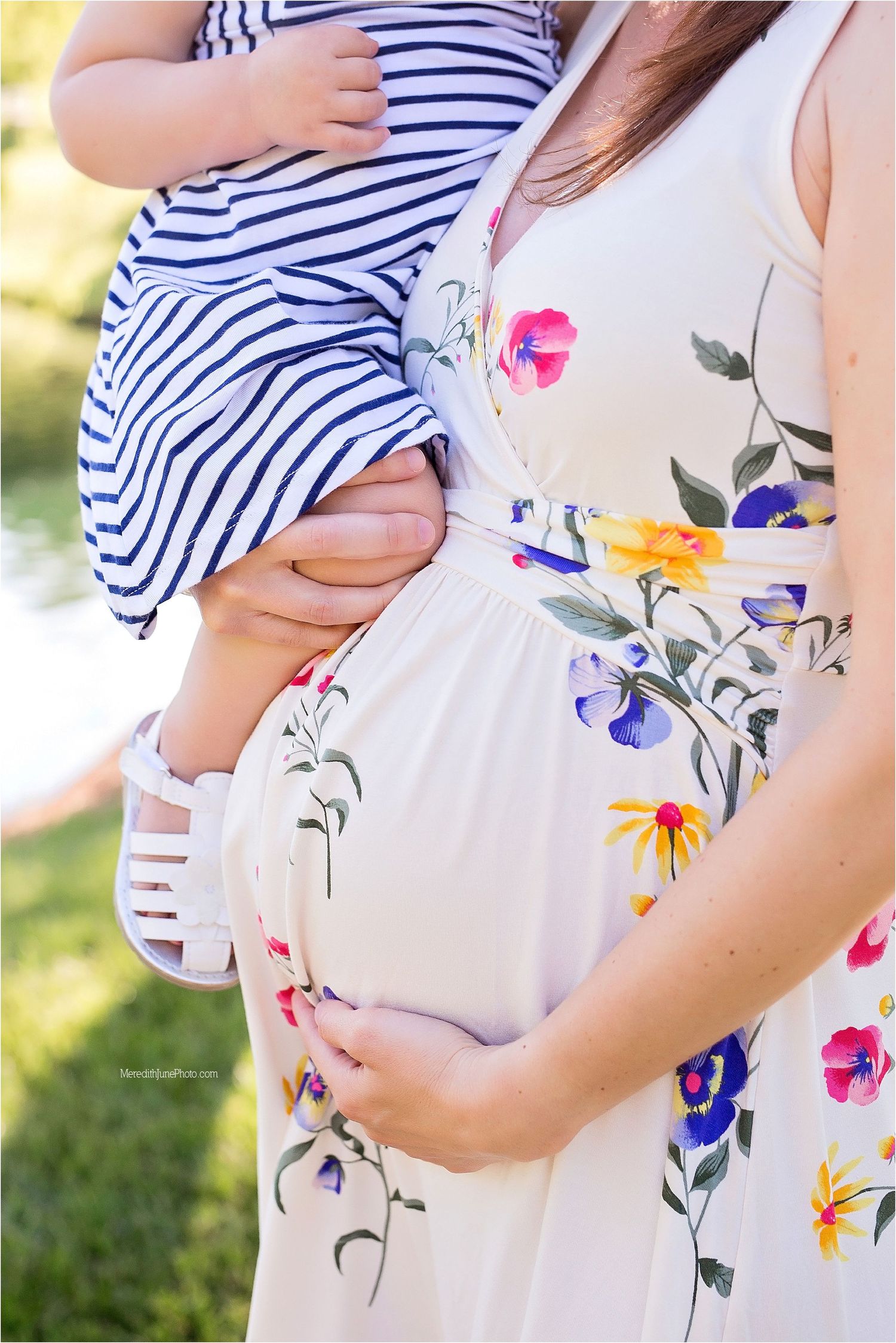 Close-up of pregnant woman in floral dress holding toddler wearing navy striped outfit in sunny outdoor setting.