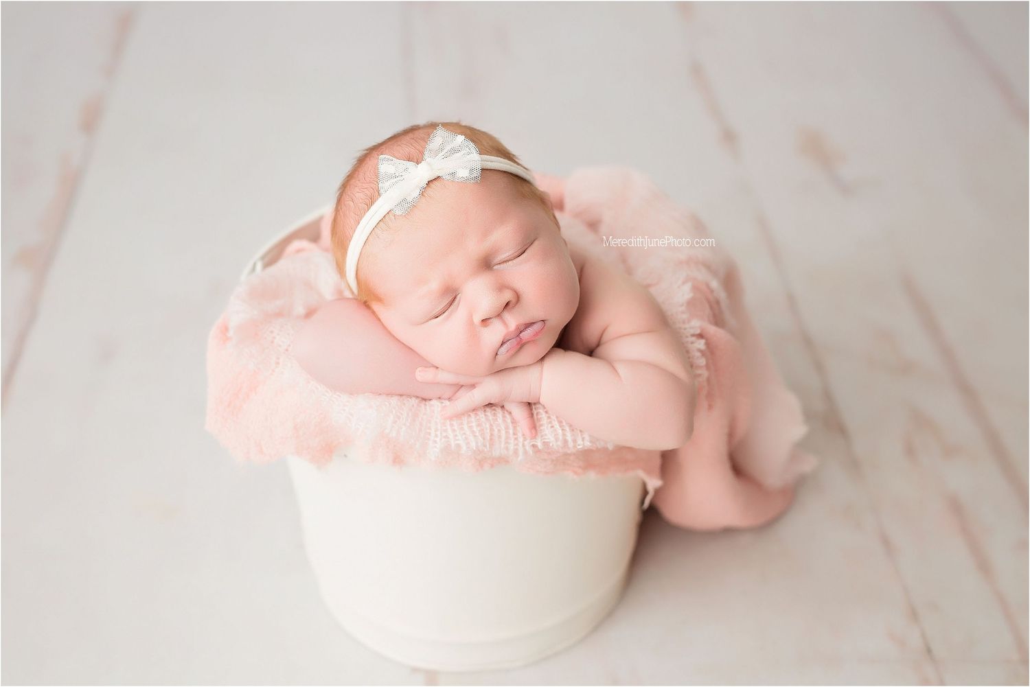 Sleeping newborn wrapped in soft pink fabric lying in white bowl against textured background.