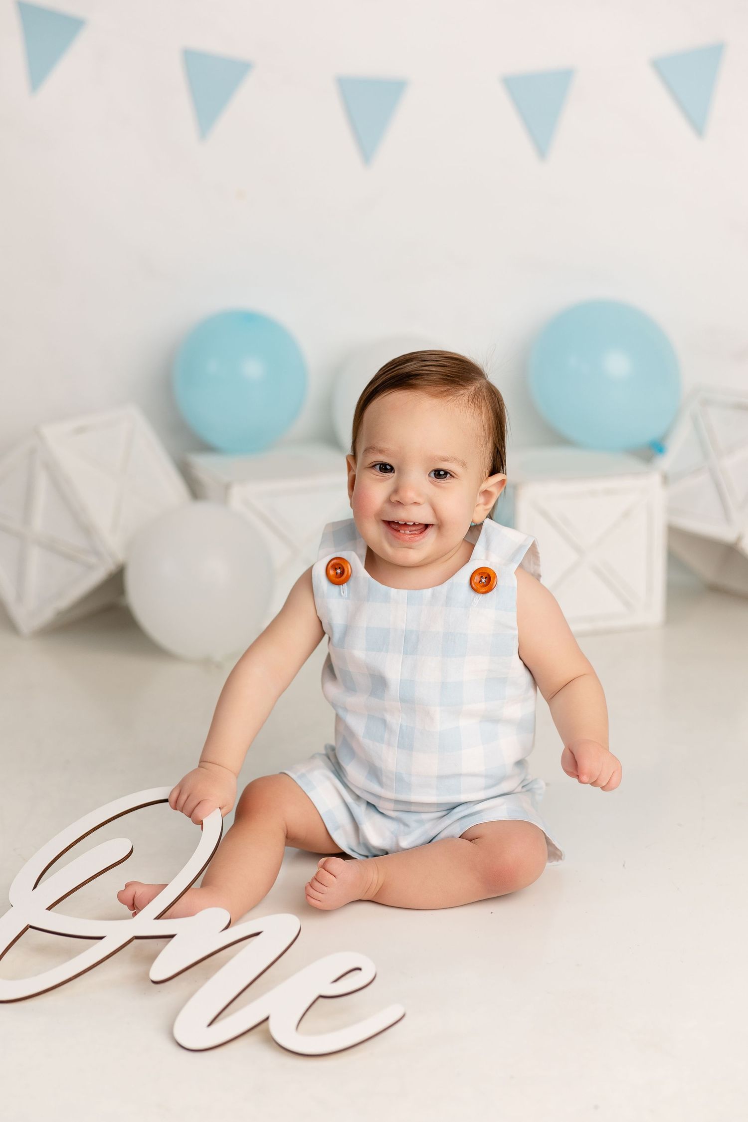 Smiling baby in blue checkered outfit sits on white floor with blue balloons and banner decorations.