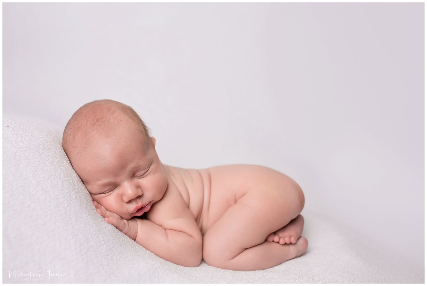 A sleeping newborn baby curled up peacefully on a soft white blanket in a professional studio setting.