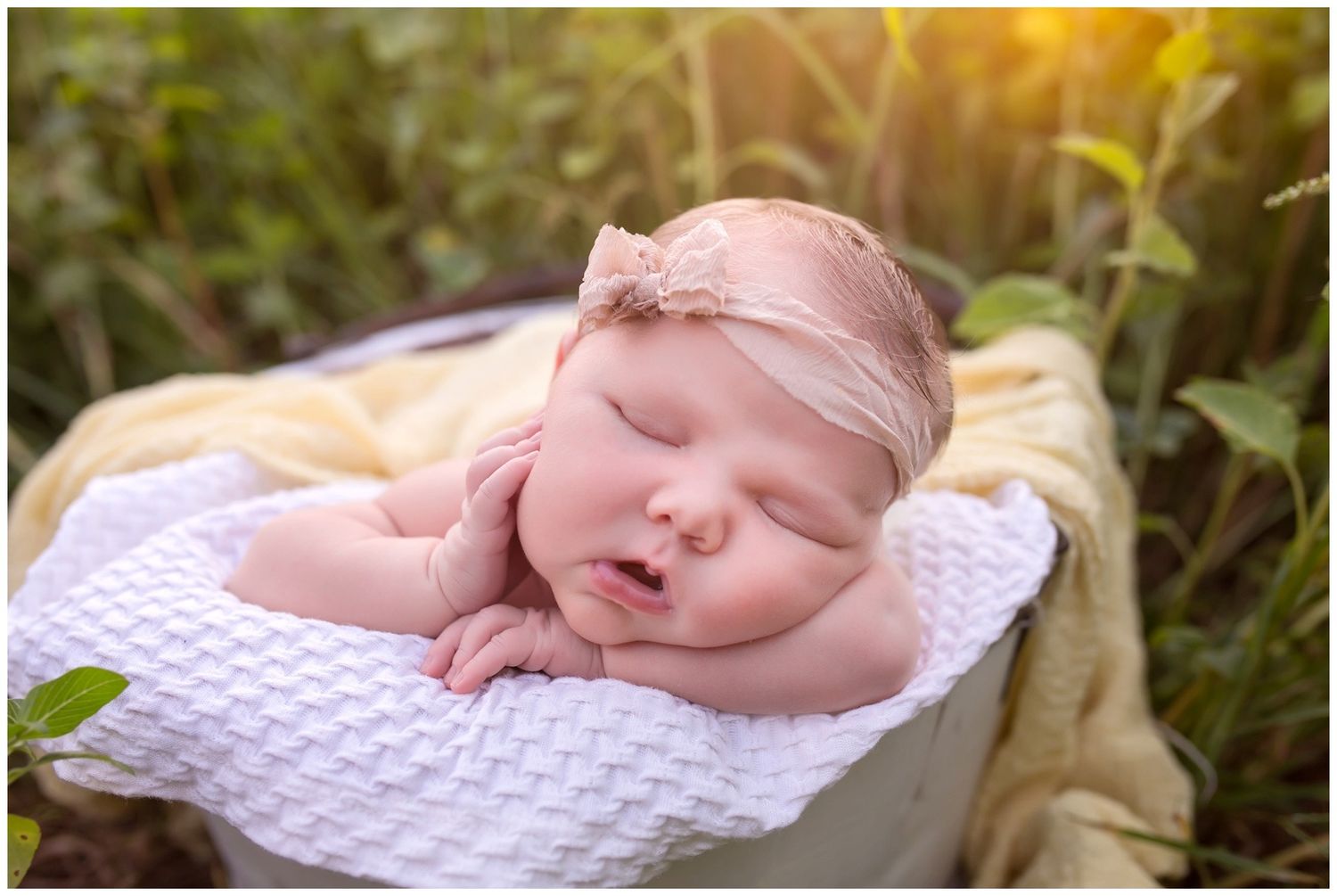 A sleeping newborn baby rests peacefully on a white textured blanket in an outdoor garden setting.