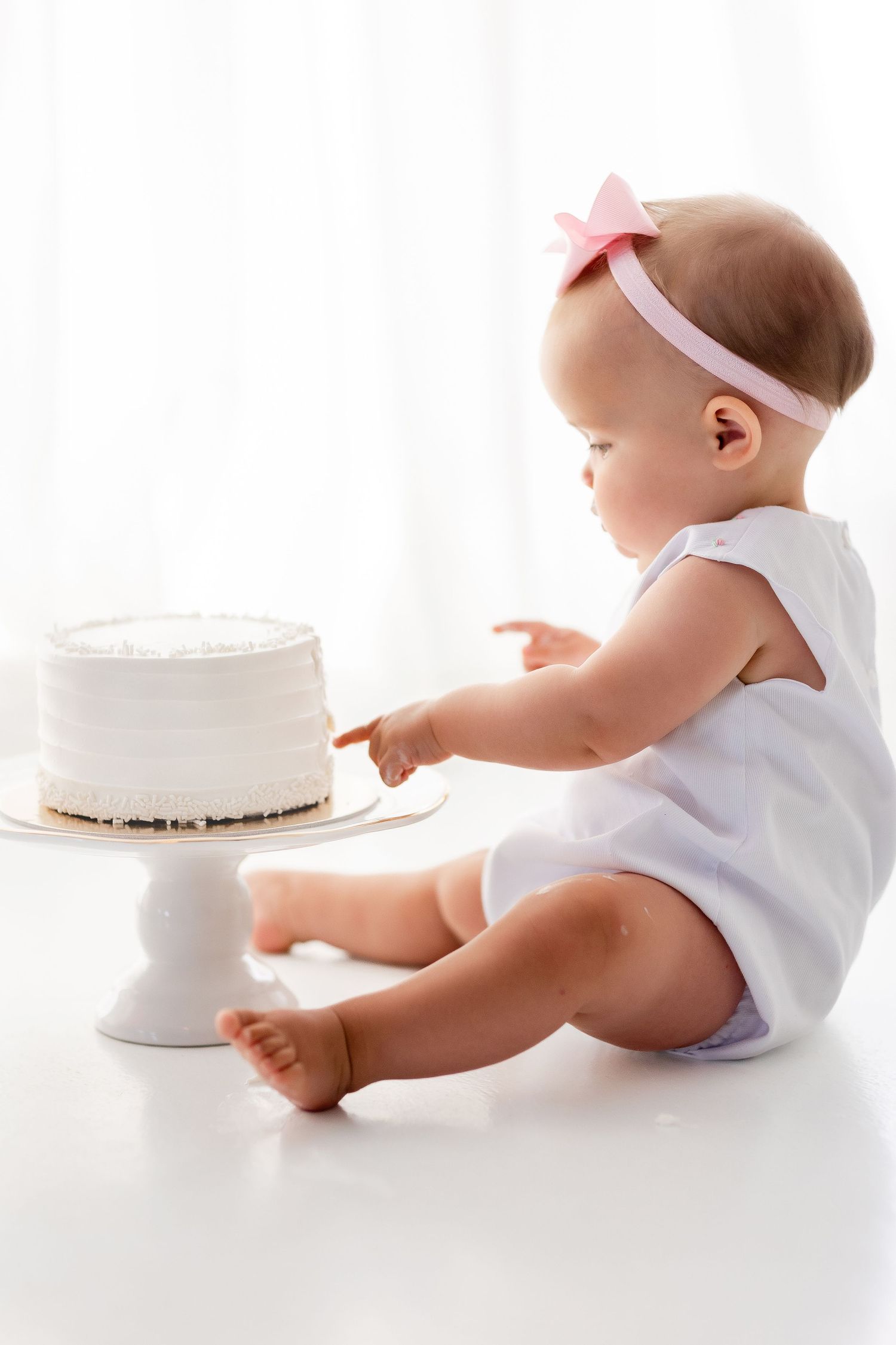 Baby in white romper and pink bow reaching towards white cake