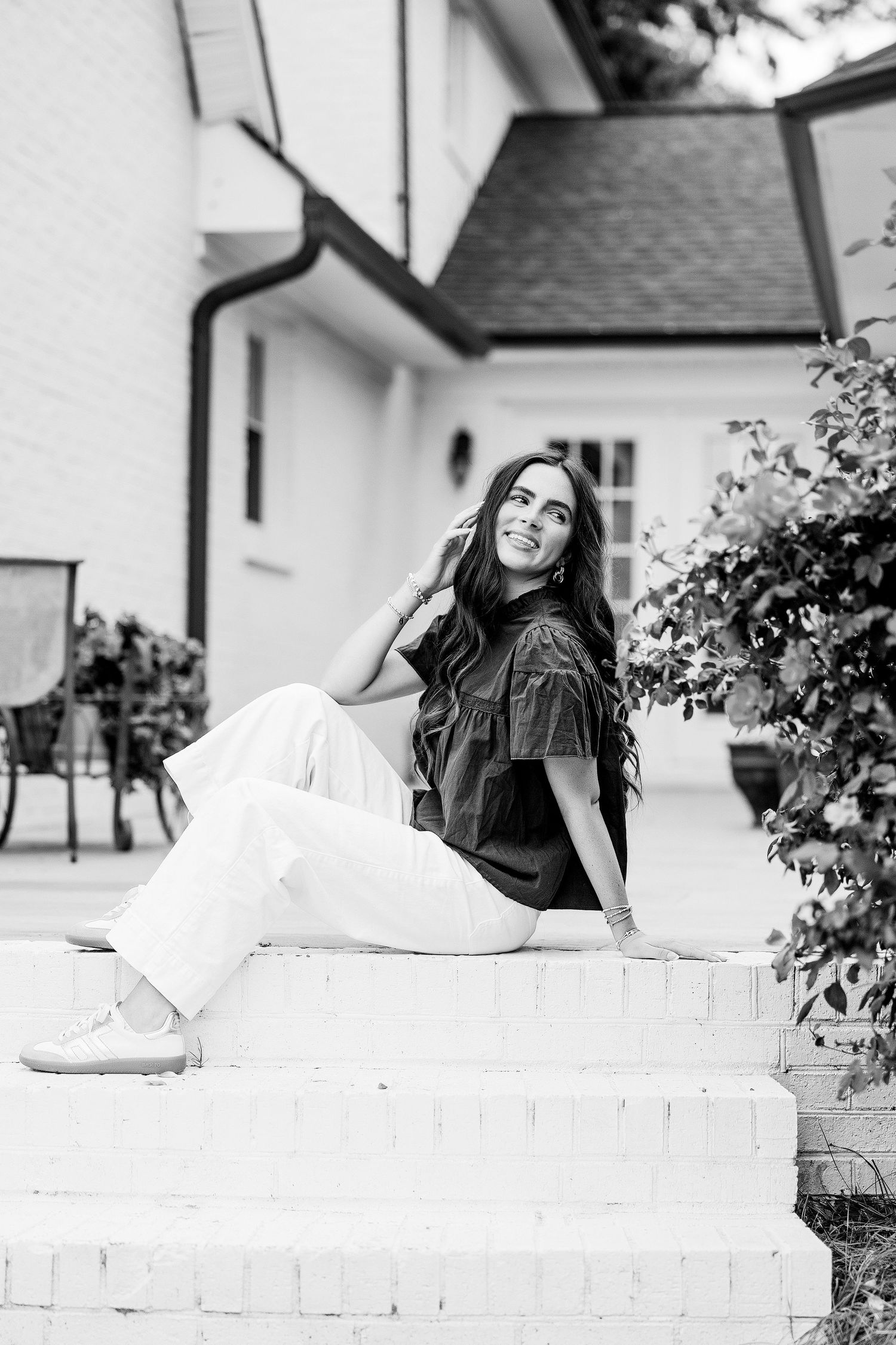 Black and white photo of a young woman with long hair sitting on white brick steps outside a house.