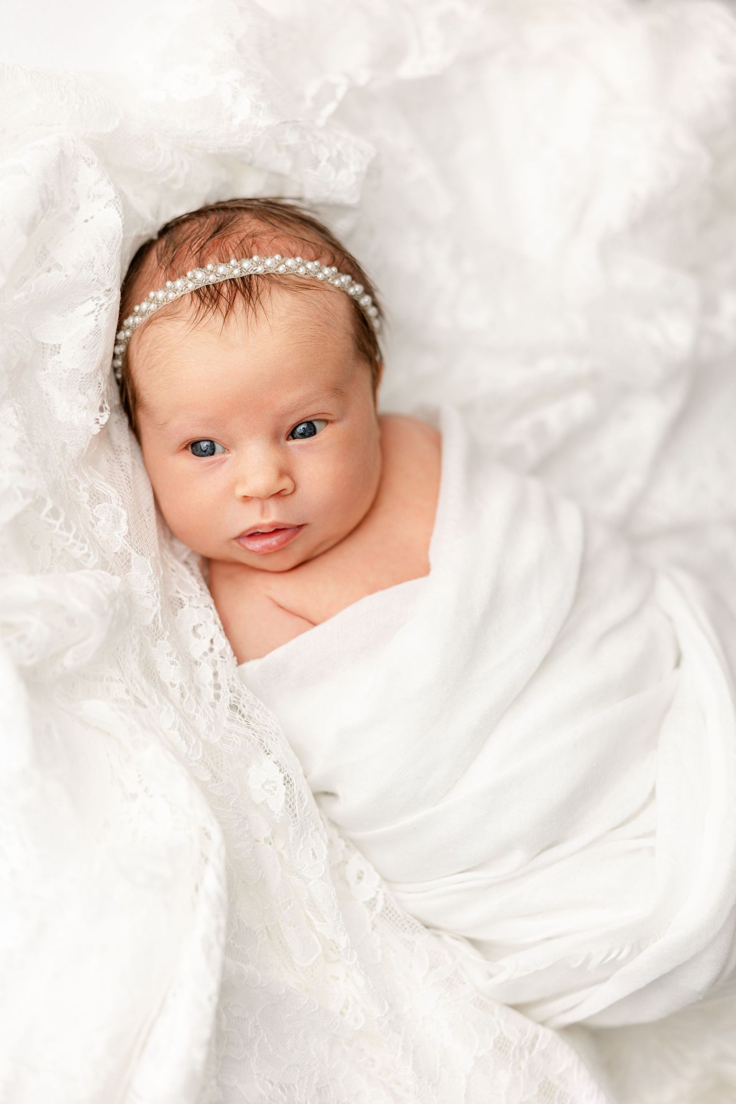 A newborn baby wrapped in soft white lace fabric gazes at the camera with bright eyes.