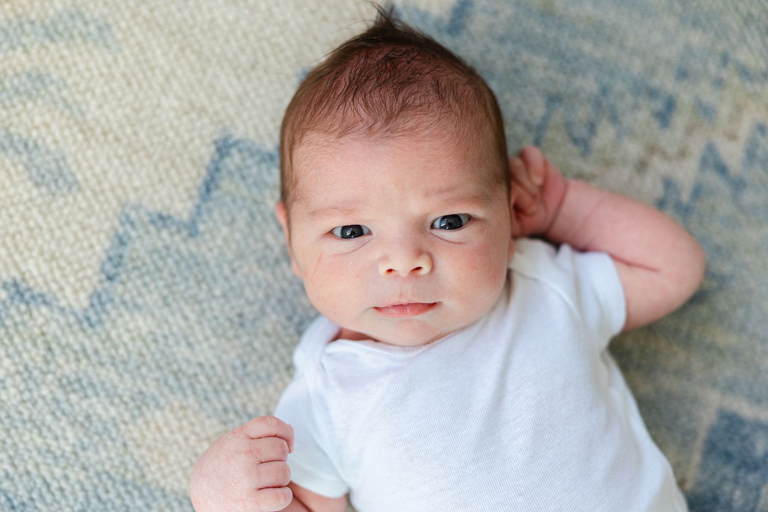 A baby wearing a white onesie lies on a textured blue blanket while looking at the camera with a gentle expression.