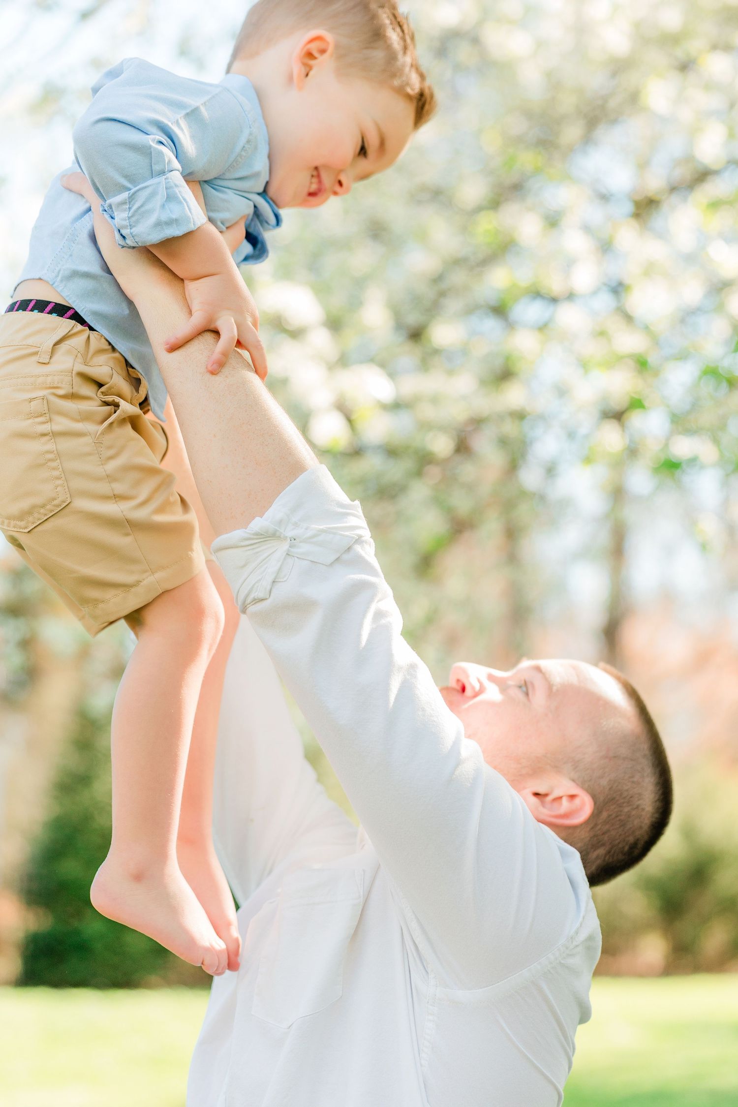 A tender moment captured outdoors as a parent lifts and plays with a small child wearing khaki shorts in spring.