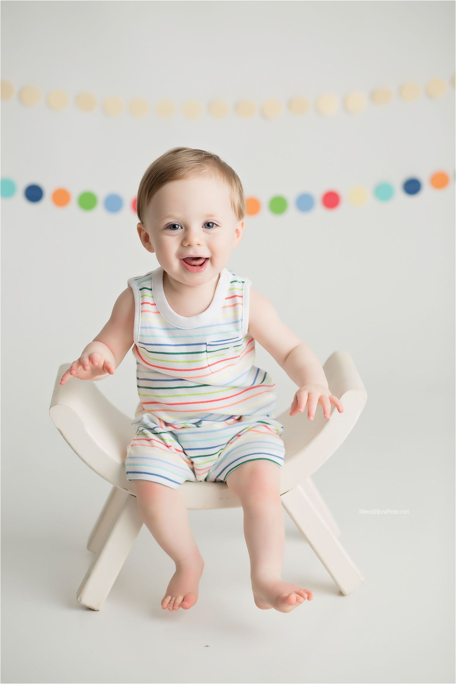 A happy baby wearing a striped romper sits in a white chair under colorful party decorations.