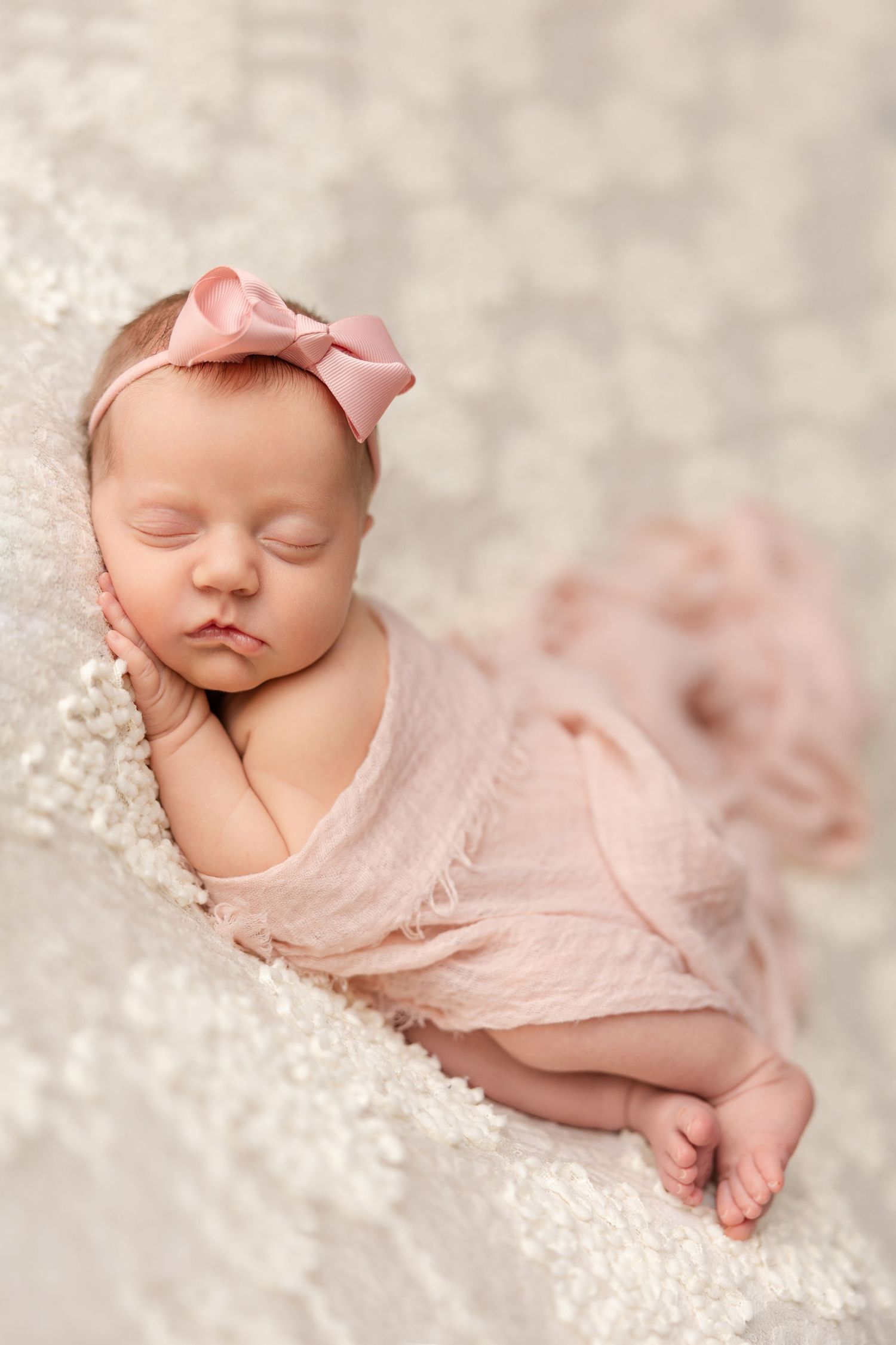 Sleeping newborn baby wearing pink bow and tulle wrap posed on soft cream textured blanket in studio setting.