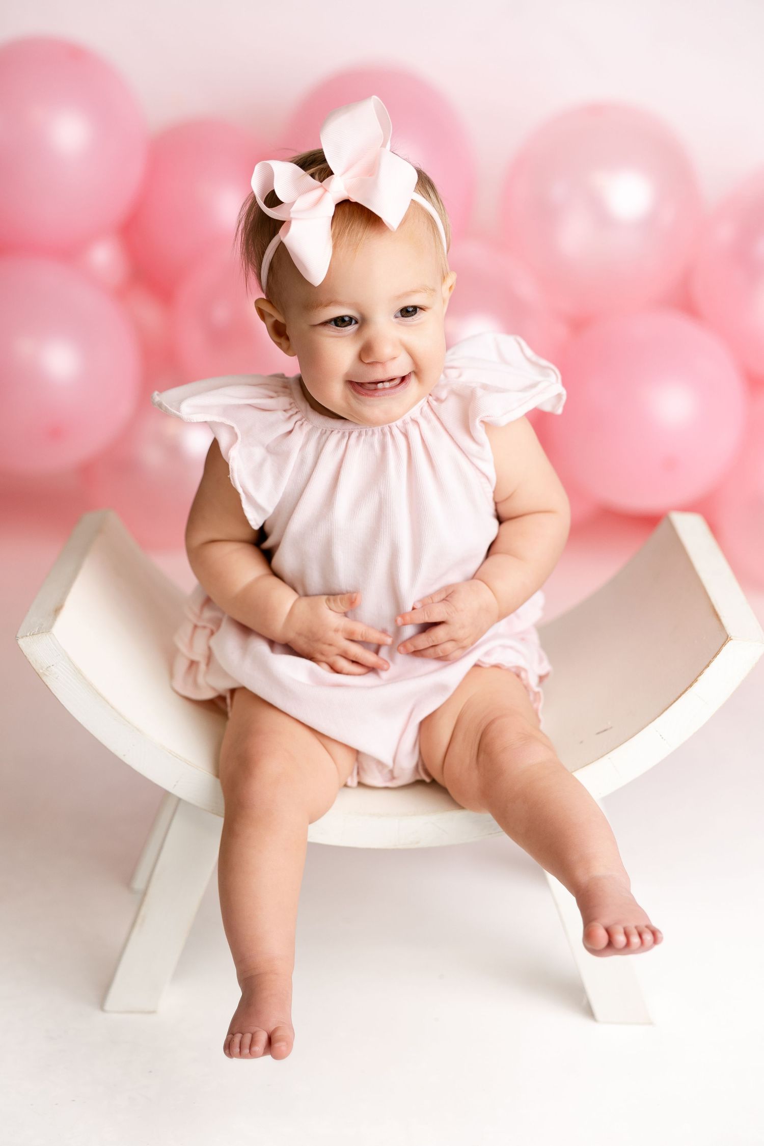 Sweet baby portrait with pink balloons wearing white dress and bow sitting on white chair.
