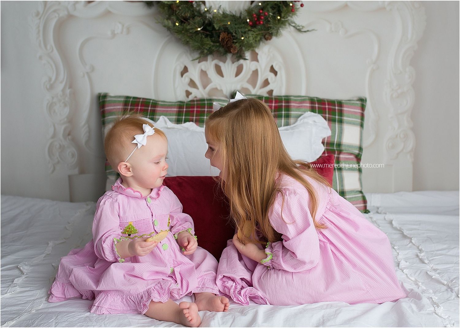 Panoramic holiday bedroom scene with plaid pillows, Christmas wreath, and pink clothing against white bedding.