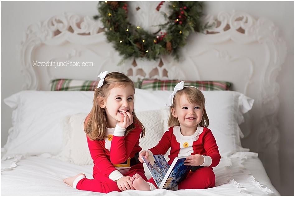 Two children in matching red pajamas sit on a white bed beneath a Christmas wreath, sharing a joyful holiday moment.