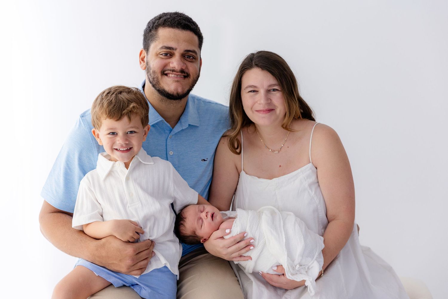 Happy family of four posing for a newborn photo session, parents smiling with toddler son and swaddled baby on white background.