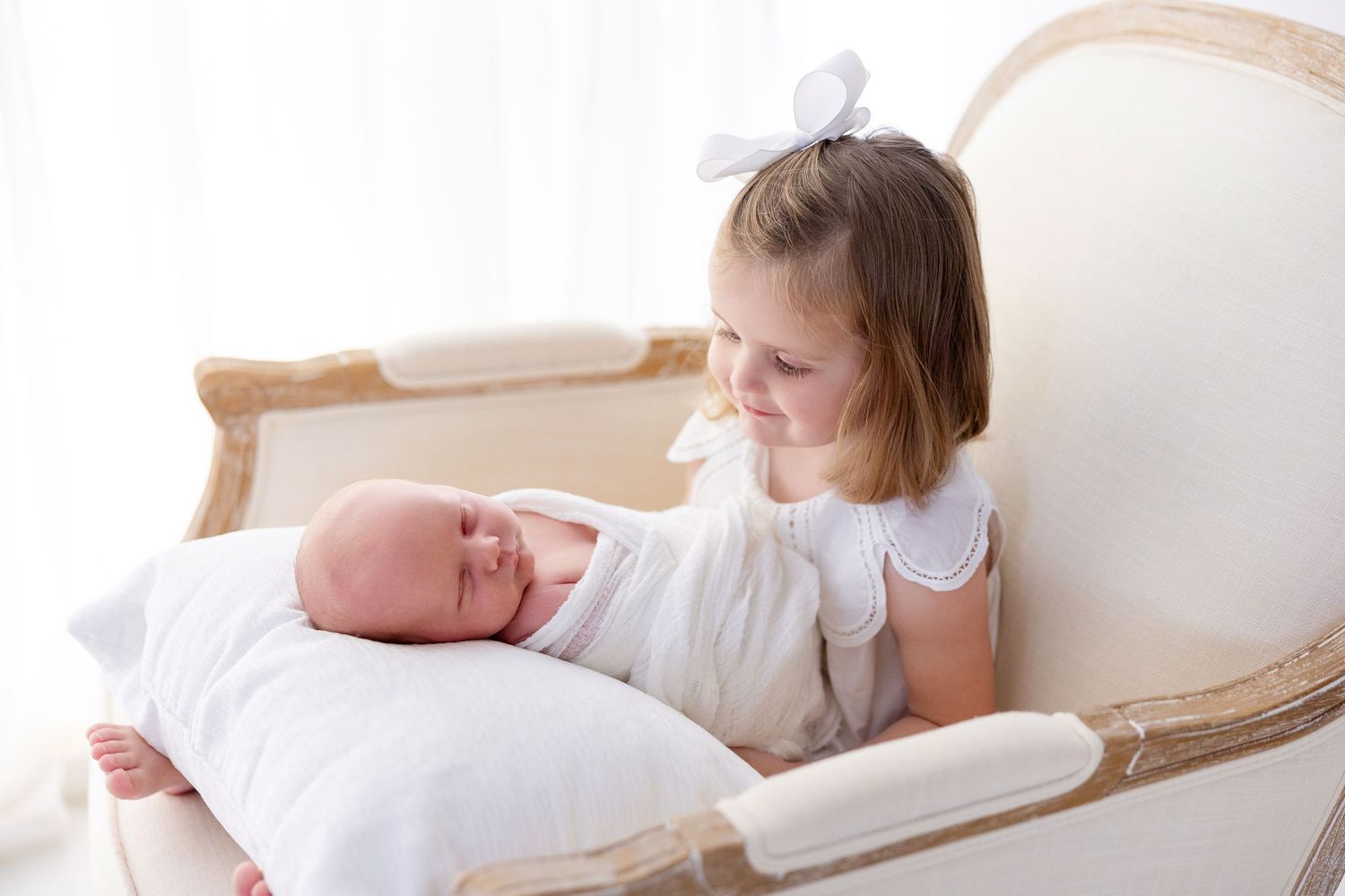 Young girl in white dress with bow sits in elegant chair holding sleeping newborn baby sibling on a pillow.