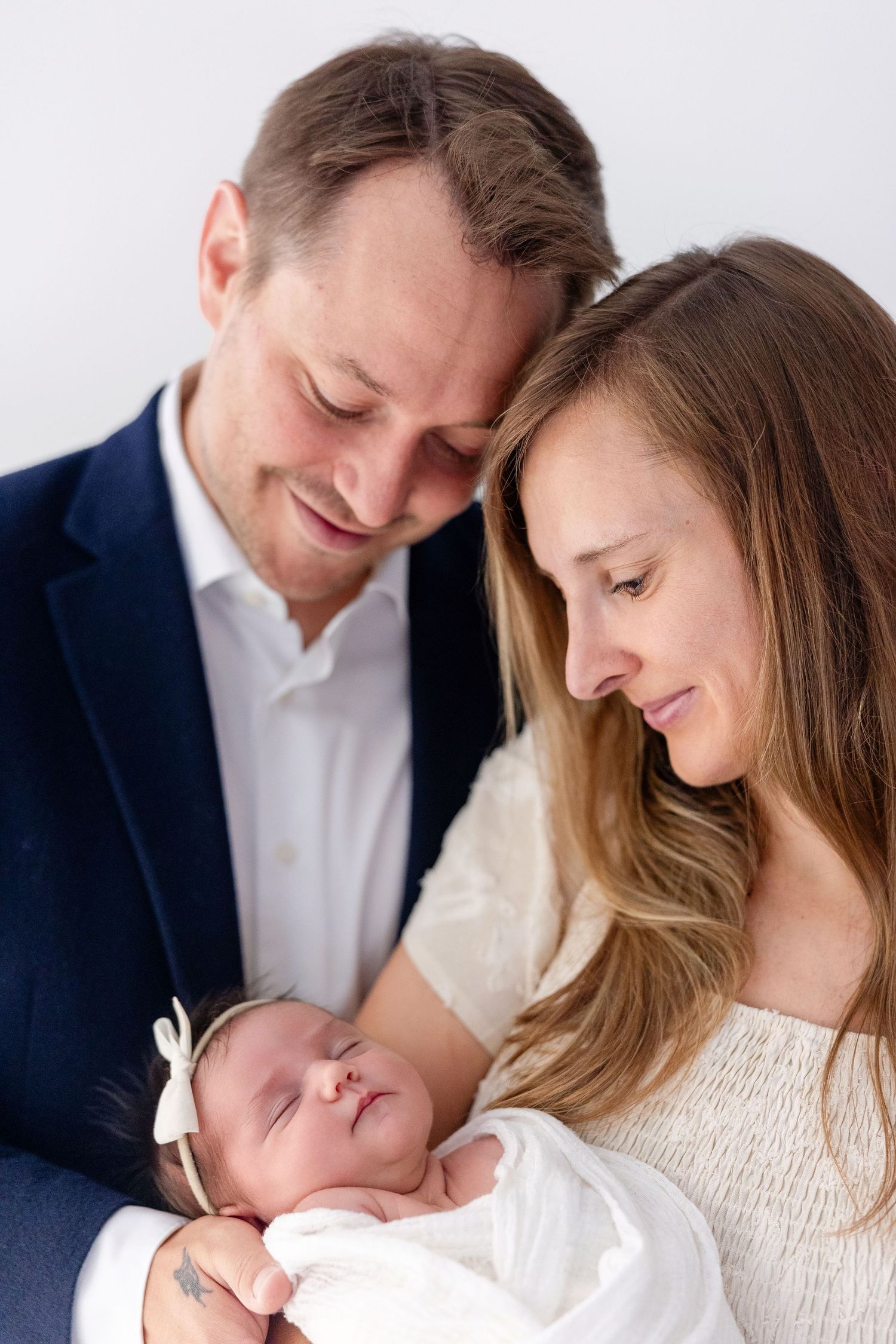Smiling parents gaze lovingly at their sleeping newborn baby wrapped in a white blanket and wearing a bow headband in a bright studio.