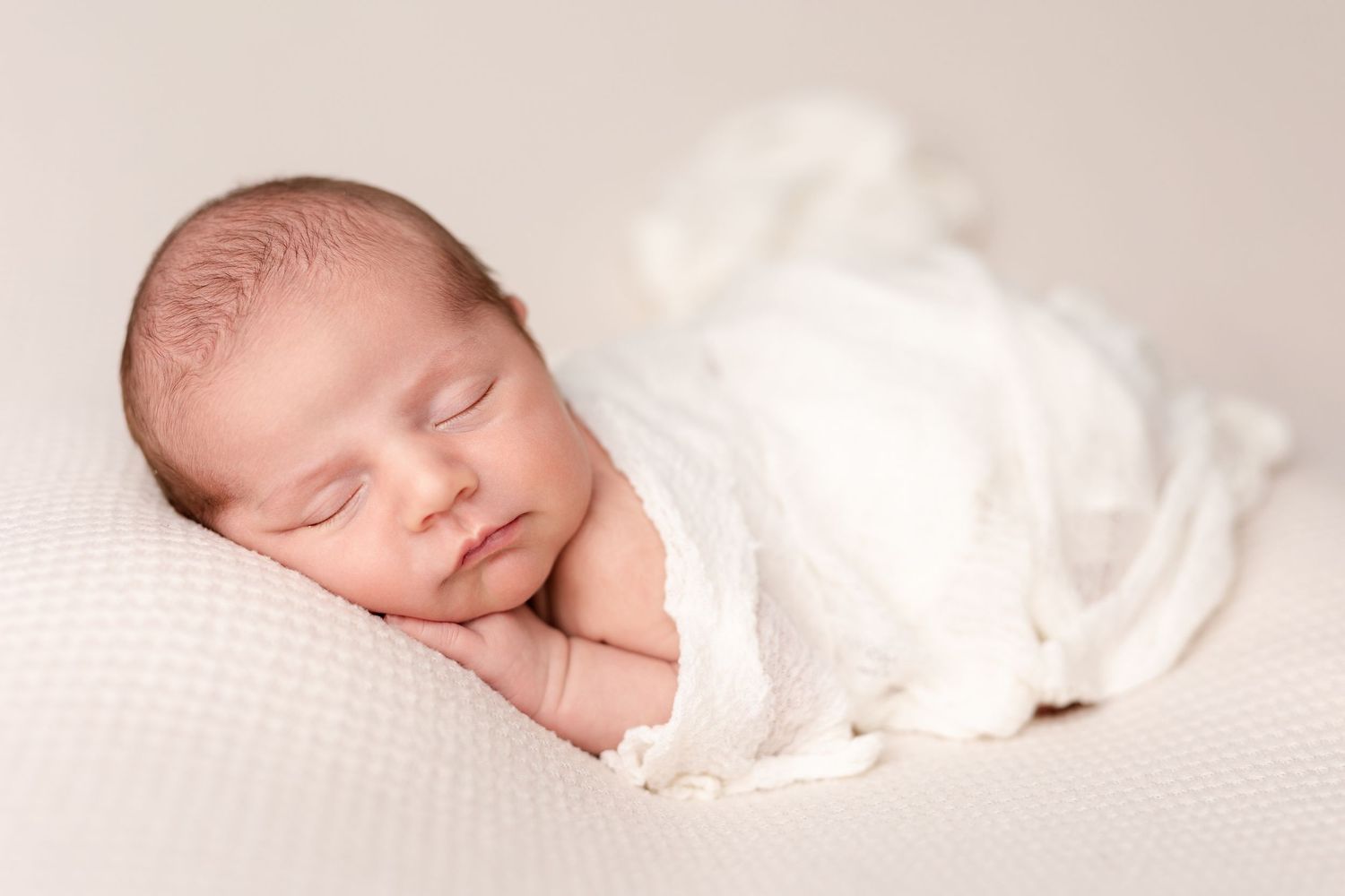 A sleeping newborn baby rests peacefully on soft white textiles in a gentle side-lying pose.
