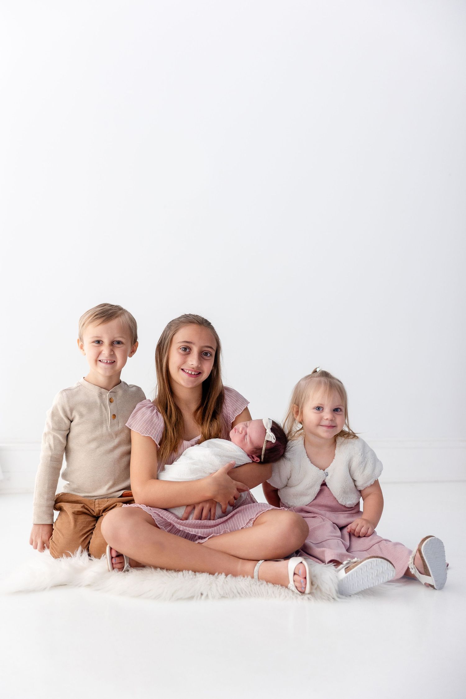 Three smiling siblings sit together on a white studio floor, with the older girl in a pink dress gently holding a sleeping newborn baby.