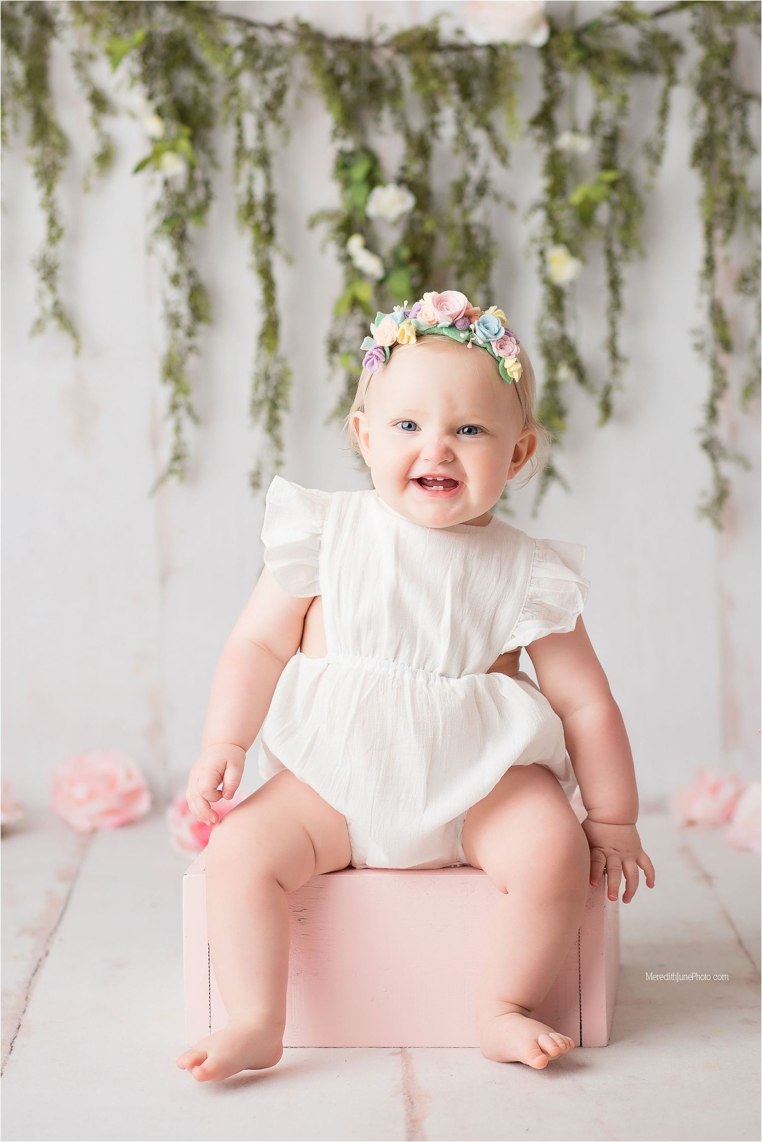 Baby in white dress with floral crown sitting against white backdrop with hanging greenery in studio photo series.