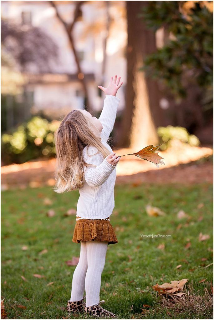 Small child in white sweater and brown skirt dances joyfully in autumn leaves at sunset.
