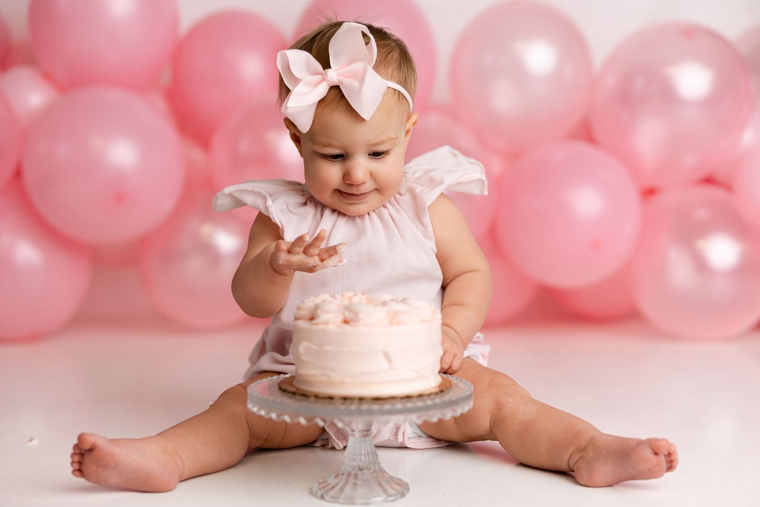 Baby in white dress sits with smash cake on pedestal against pink balloon backdrop during first birthday photoshoot.