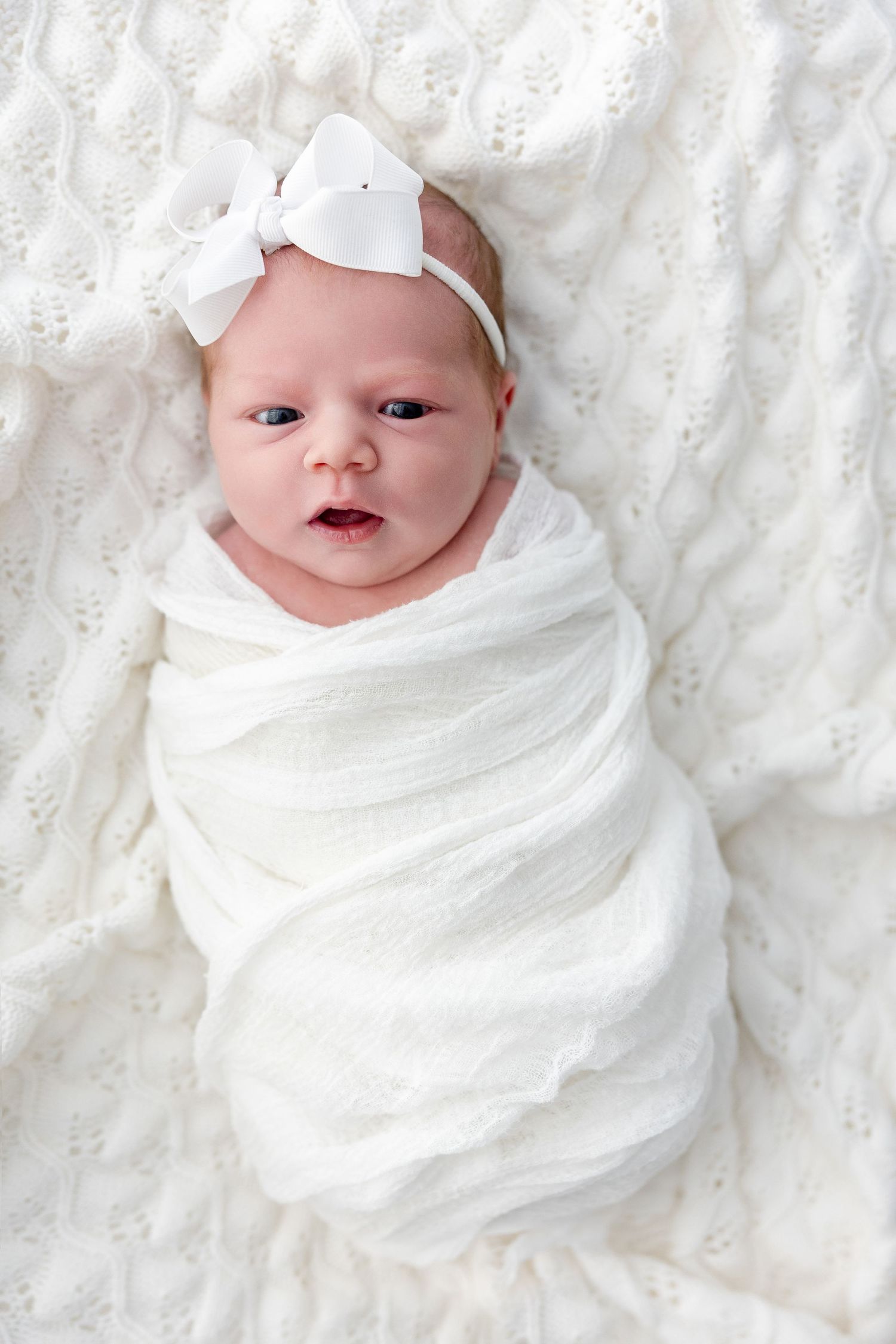 A tiny baby swaddled in white fabric with a white bow headband lies on a textured white blanket.