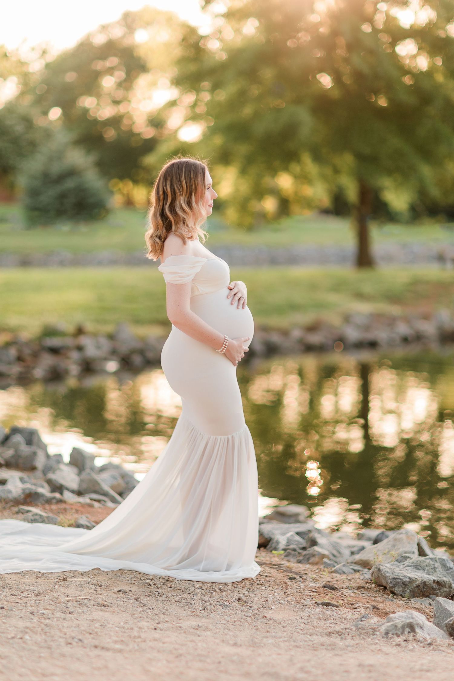 Maternity photos at sunset by rocky waterfront in flowing white dress.