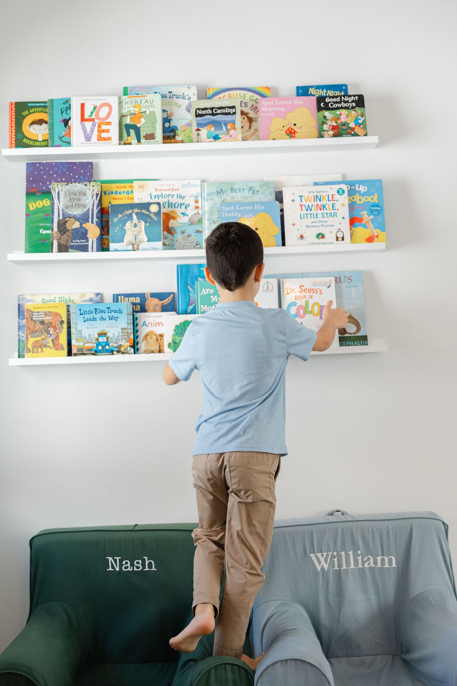 Children reaching for books displayed on white floating shelves mounted on a wall in a reading nook.