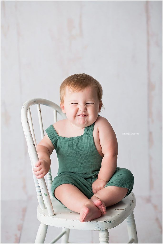 A baby wearing a green romper sits and smiles while posing on a white vintage chair in a light studio setting.