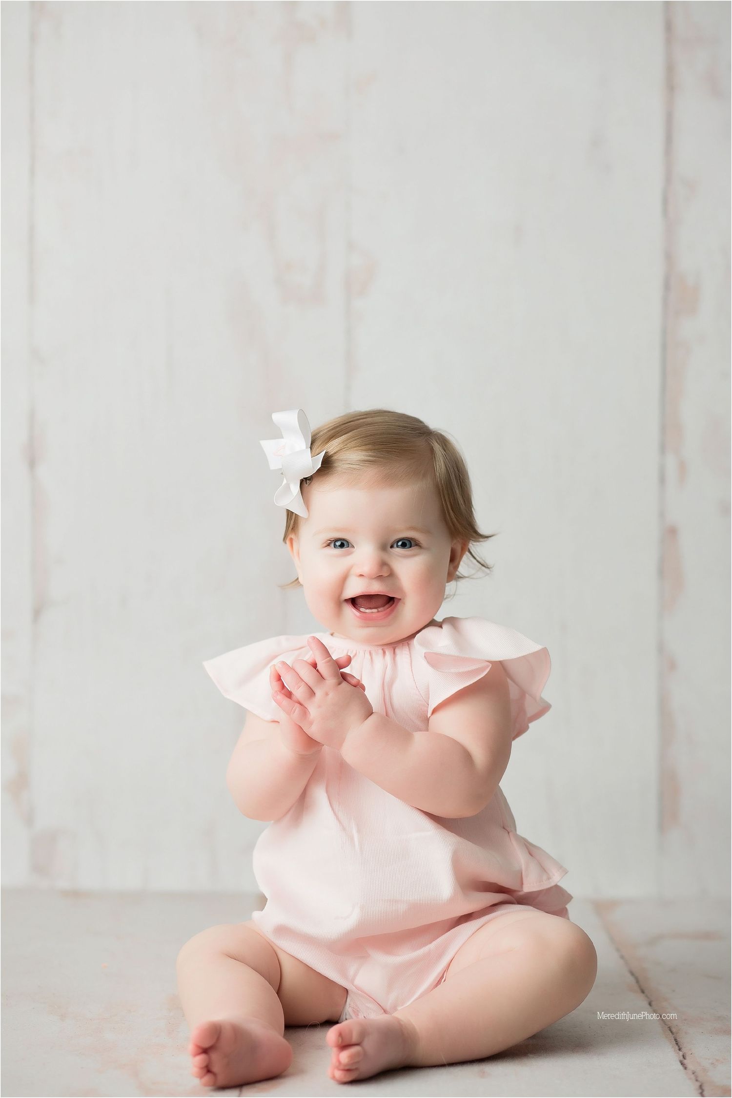 Baby in pink ruffled outfit sitting on white floor during first birthday photoshoot with soft lighting.