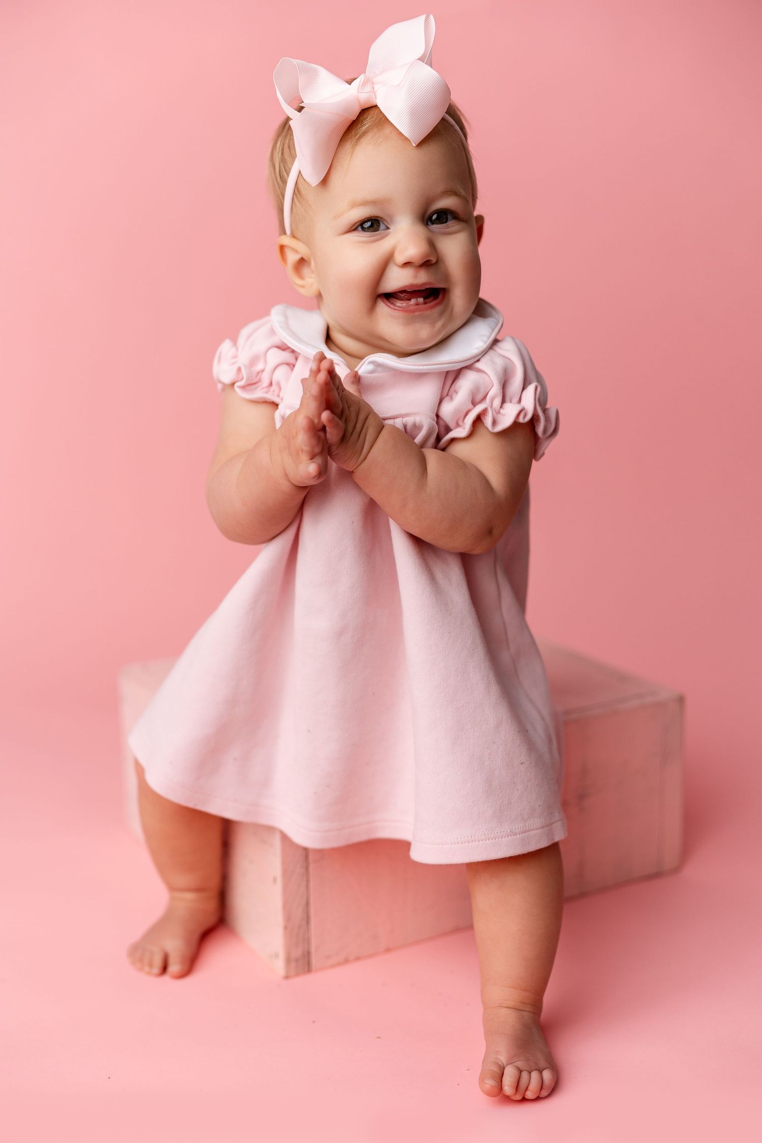Baby in pink dress sitting and smiling against pink background in studio portrait session.