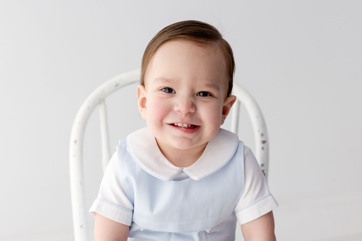 A small child in a white shirt and blue bib sits against a white background with a bright expression.