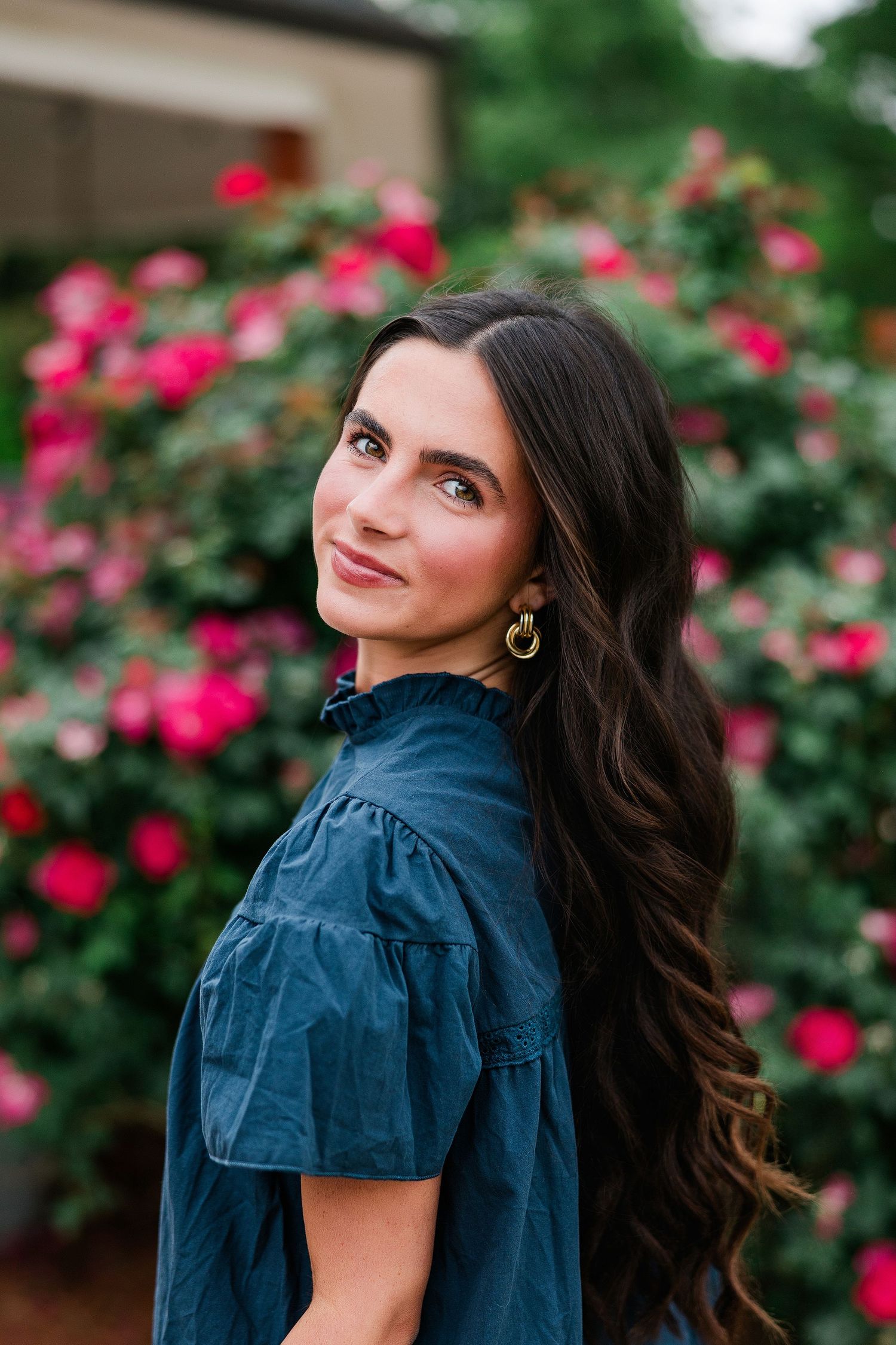 Young woman with long dark hair in a blue dress smiling outdoors near vibrant pink rose bushes.