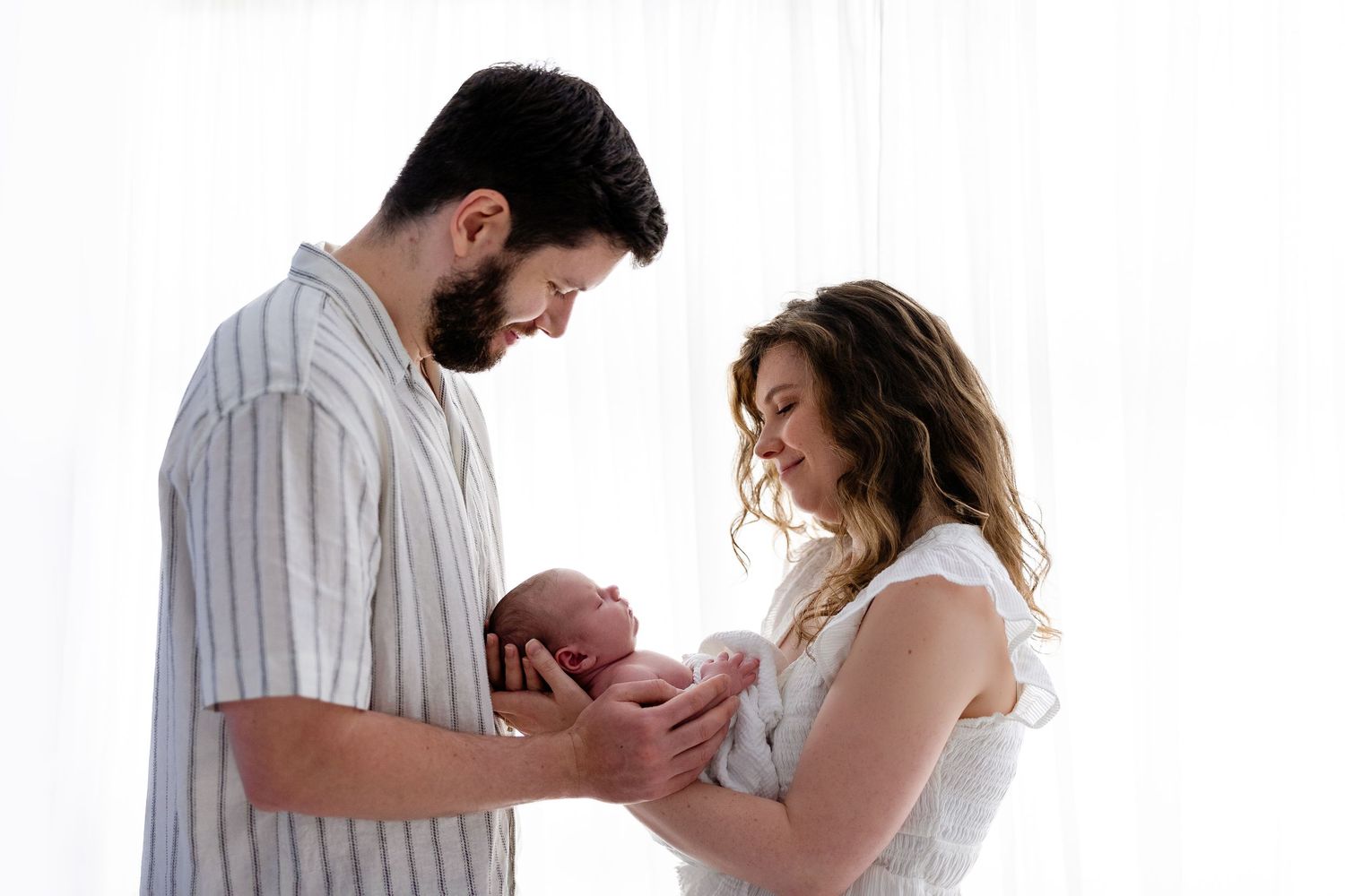 New parents tenderly holding their newborn baby together near a bright window in a heartwarming family portrait.
