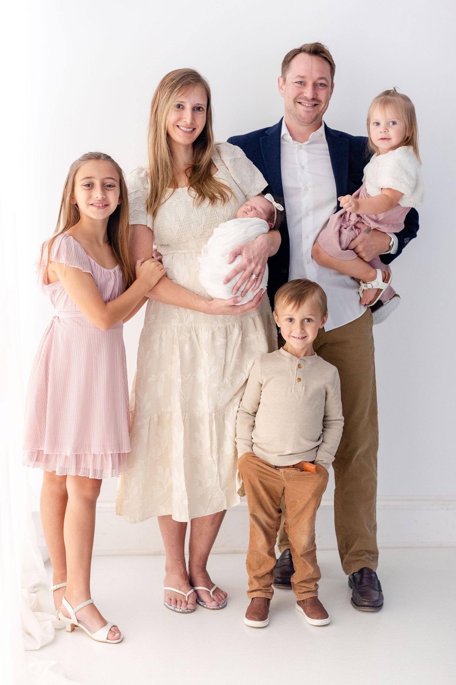 Smiling family of six in soft neutral outfits posing together against a bright white studio backdrop, with parents holding two babies.