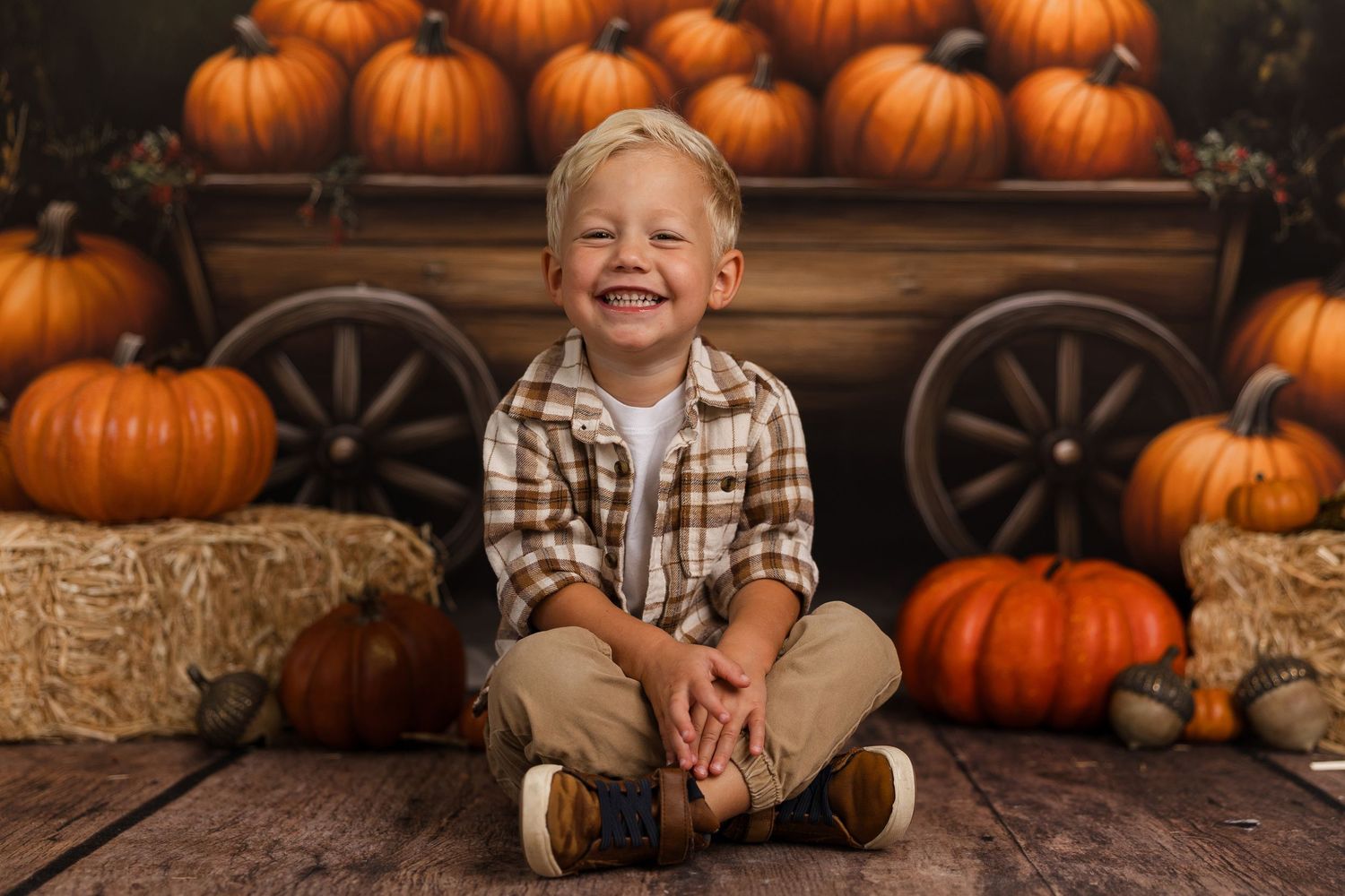 A cheerful young boy sits cross-legged on wood floor surrounded by orange pumpkins and hay bales in a fall harvest photo.