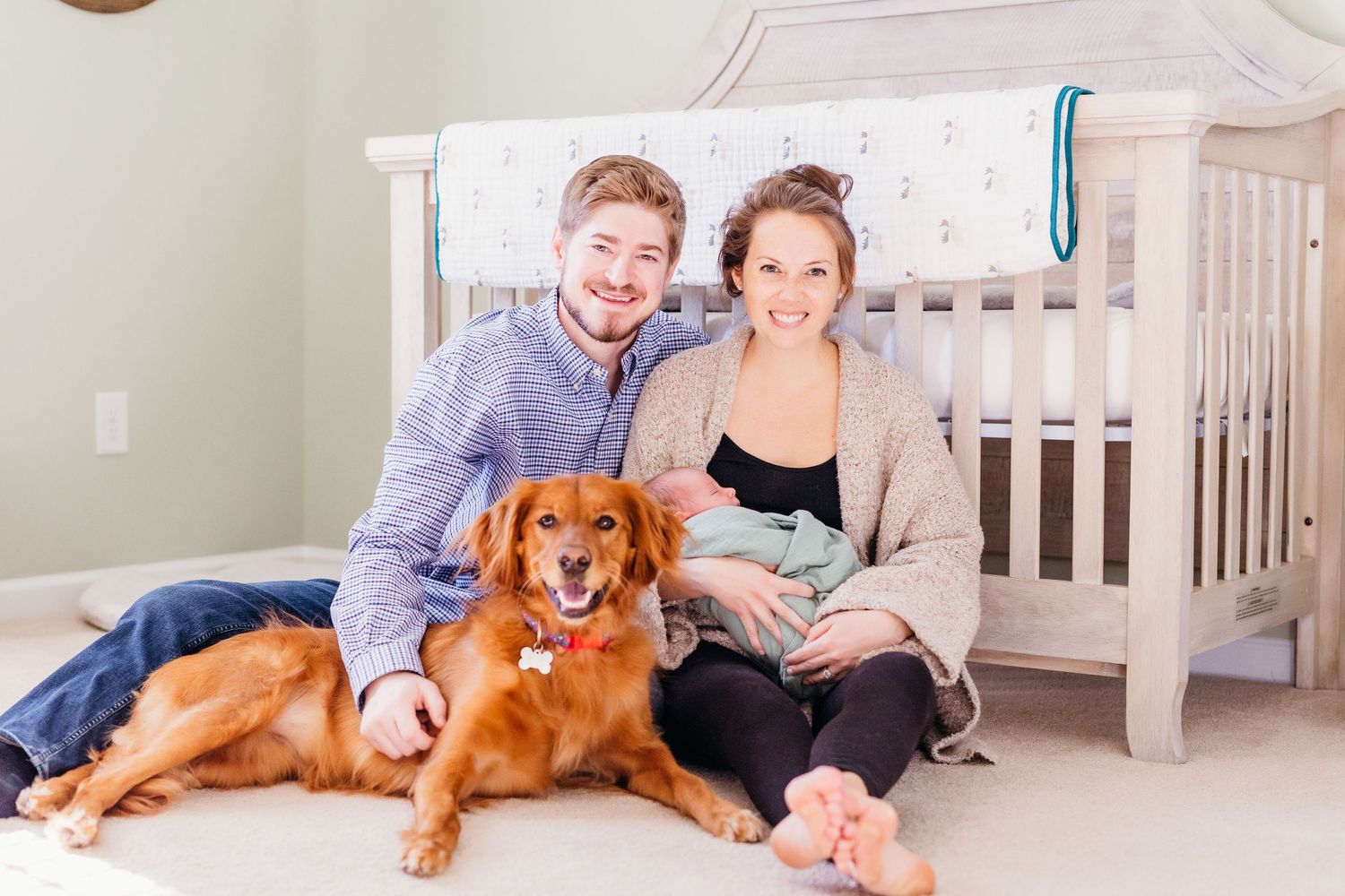A couple sits on the floor with their golden retriever dog and baby in a bright nursery room.