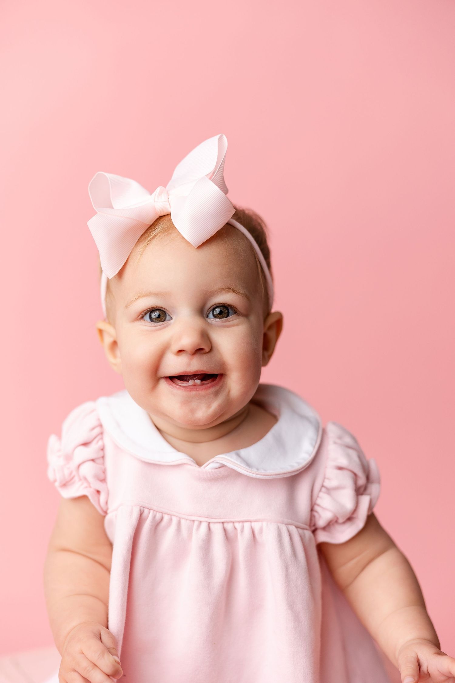 Baby wearing pink dress with white collar and bow against pink background smiles sweetly at camera.