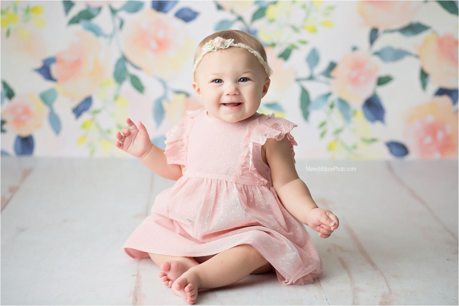 A baby in a pink dress sits and plays on a white blanket with a pastel floral backdrop.