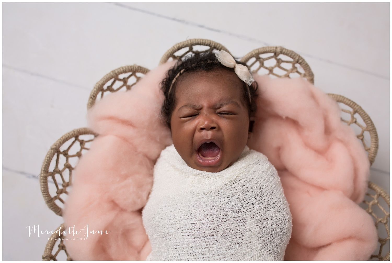 A newborn baby wrapped in white swaddling cloth lies peacefully on a soft pink blanket during a professional photoshoot.