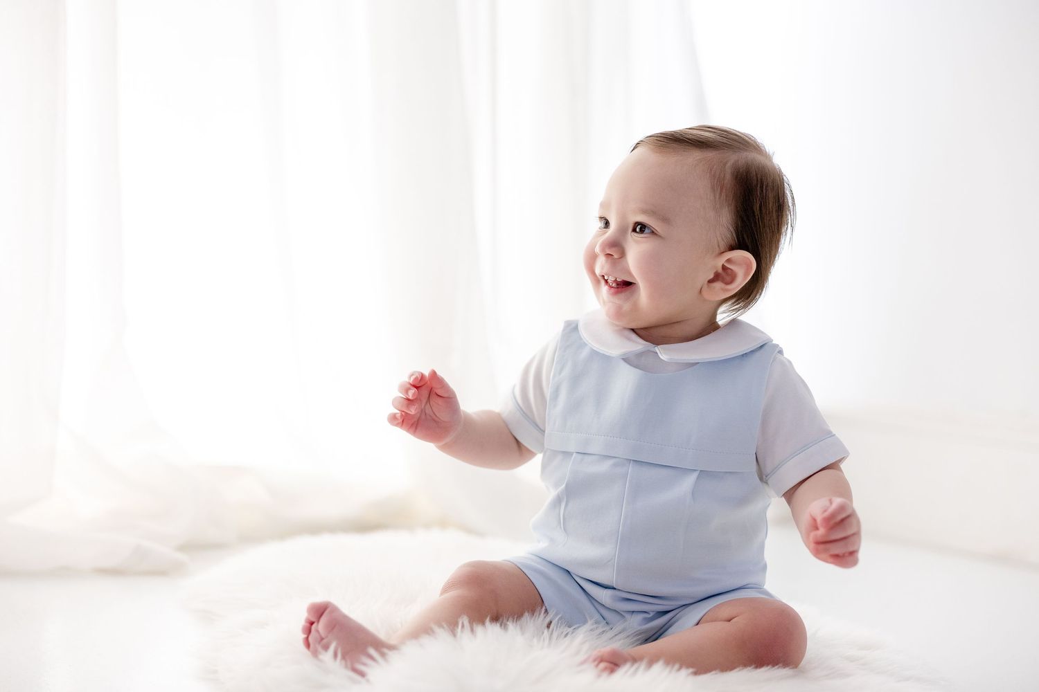 Baby wearing light blue onesie sits on white blanket in bright studio setting.