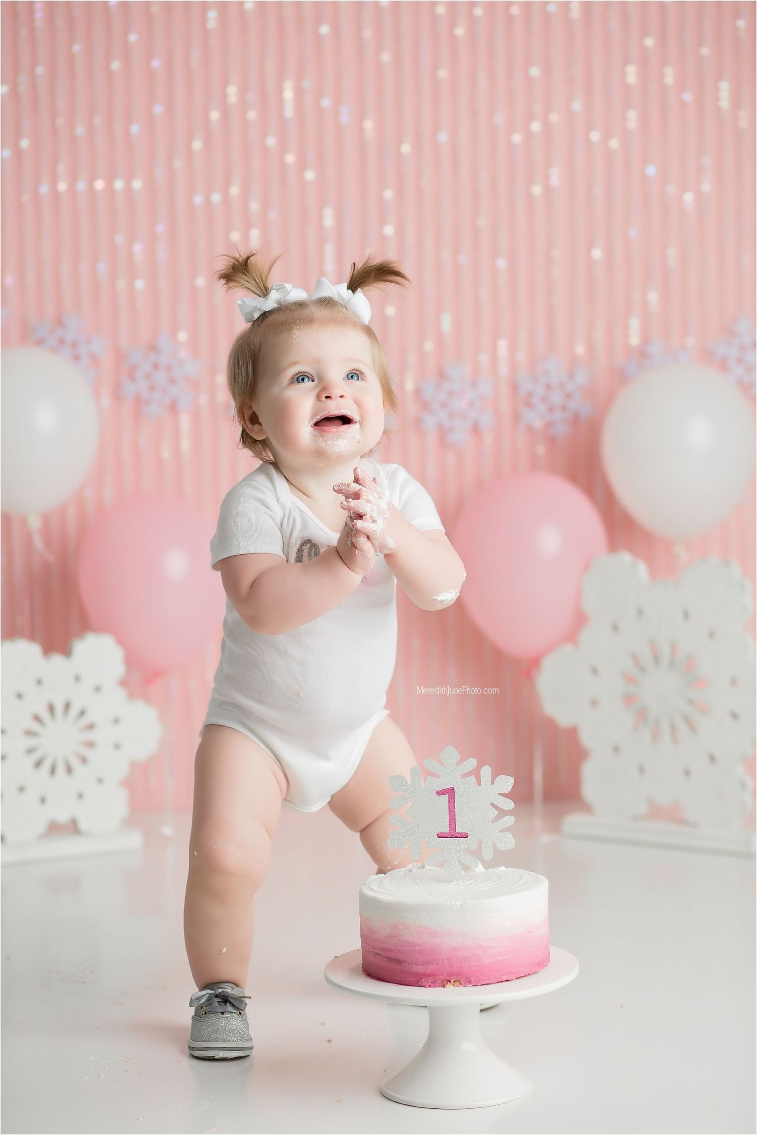 Pink and white cake smash photo session setup with balloons and decorative snowflake backdrops for first birthday celebration.